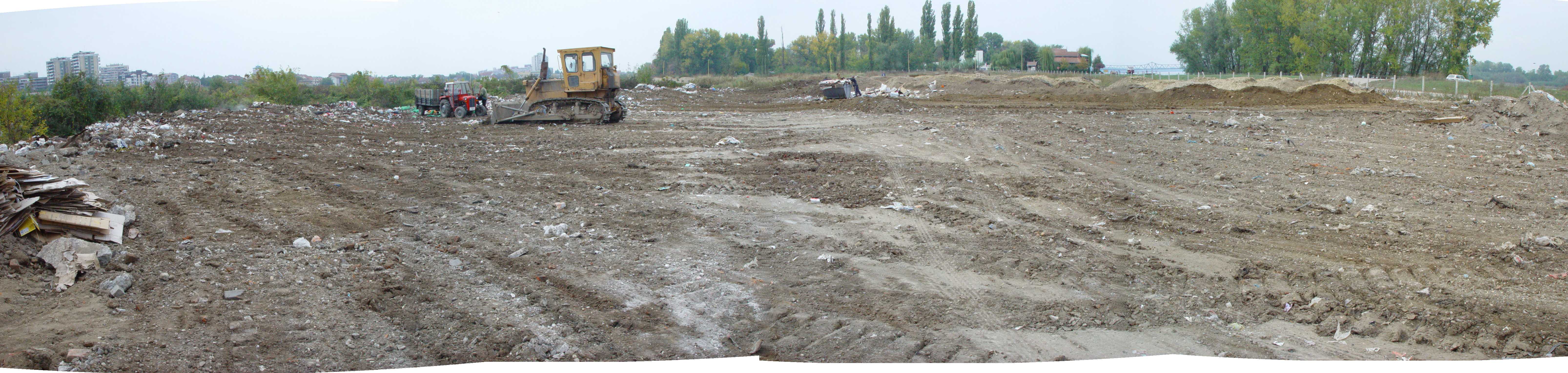 Panoramic view of a dirt lot with a bulldozer and tractor, scattered debris, trees and distant buildings under an overcast sky, analog film grain
